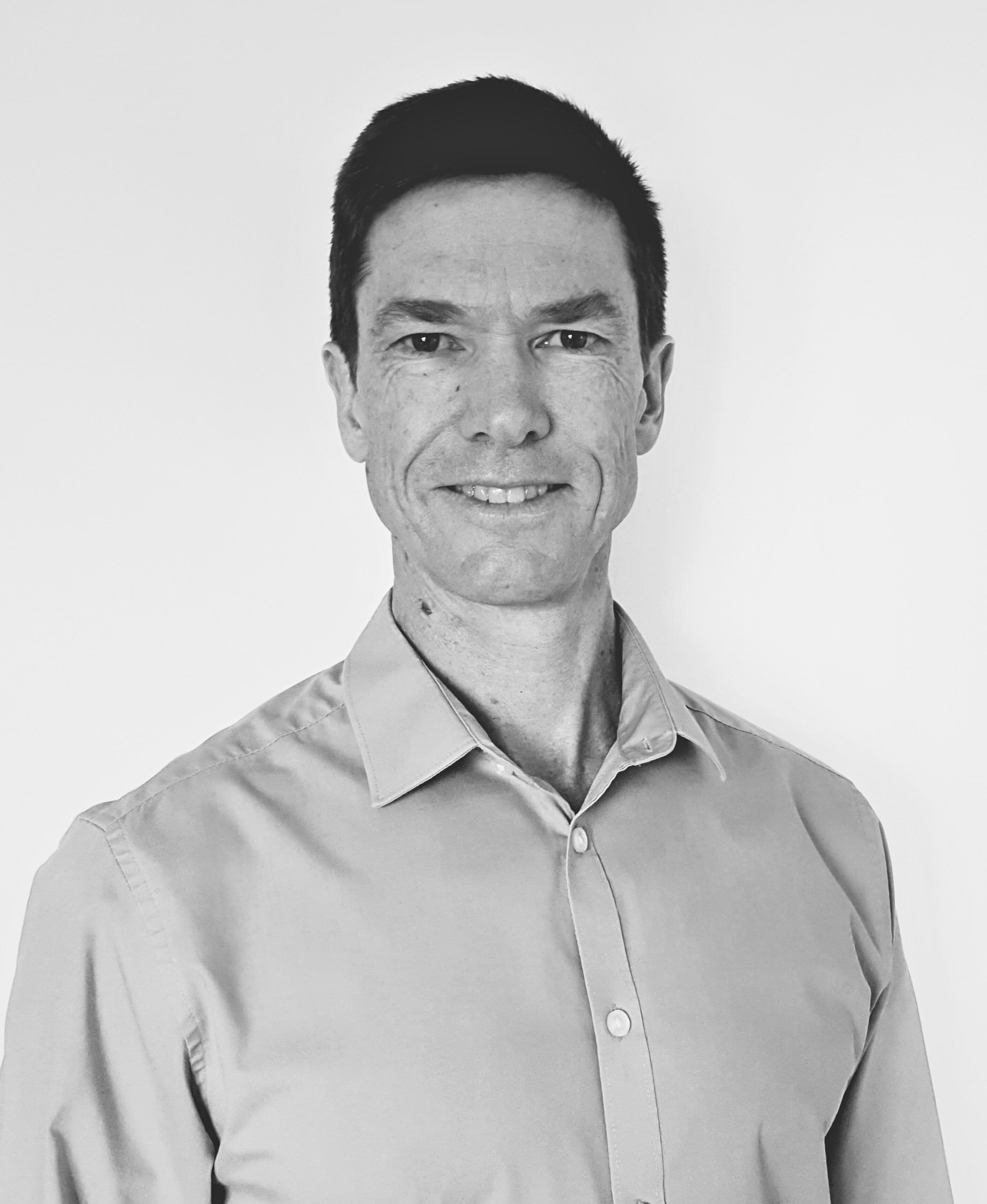 Jeremy Wakeford in a shirt stands in front of a white wall, smiling at the camera
