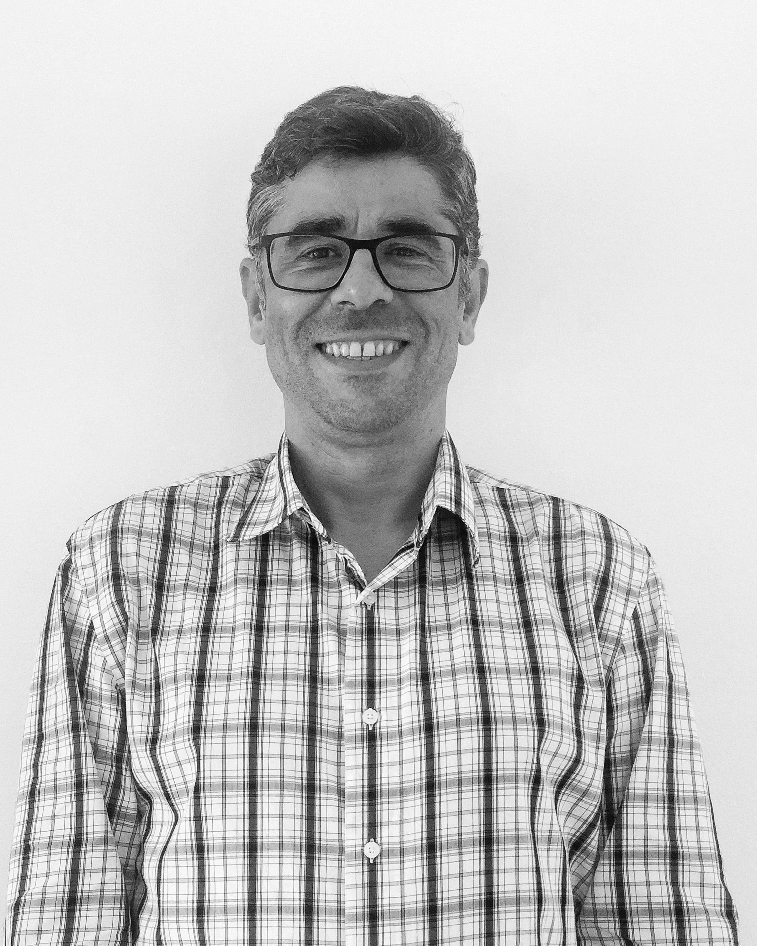 João Guimarães stands in front of a white wall, wearing a checked shirt, smiling at the camera