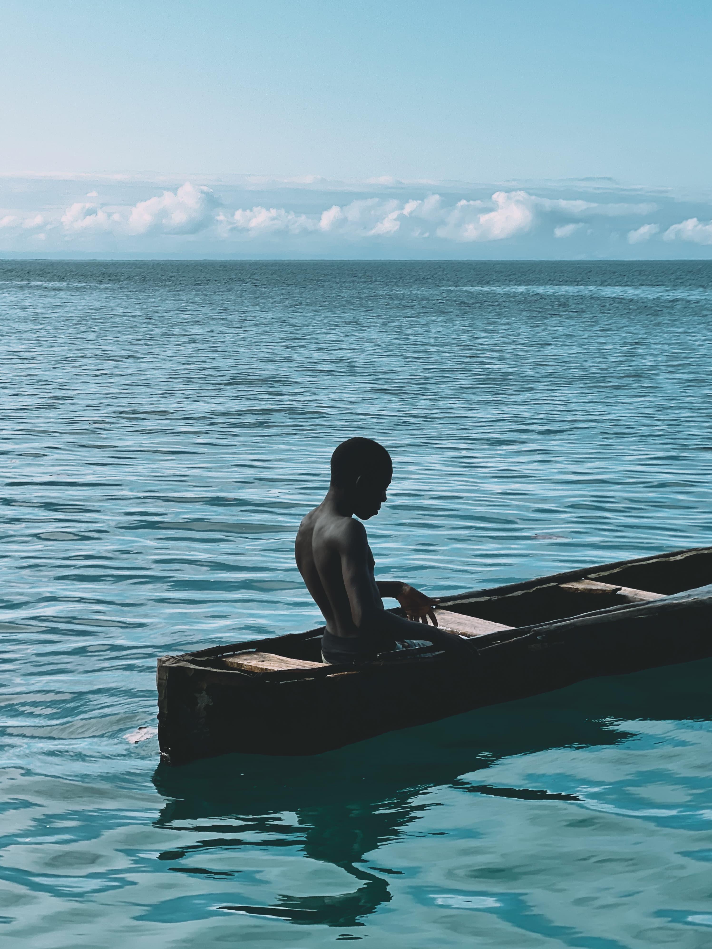 A young man sitting shirtless in a small, narrow rowing boat on the sea, looking away, seen from a diagonal rear angle