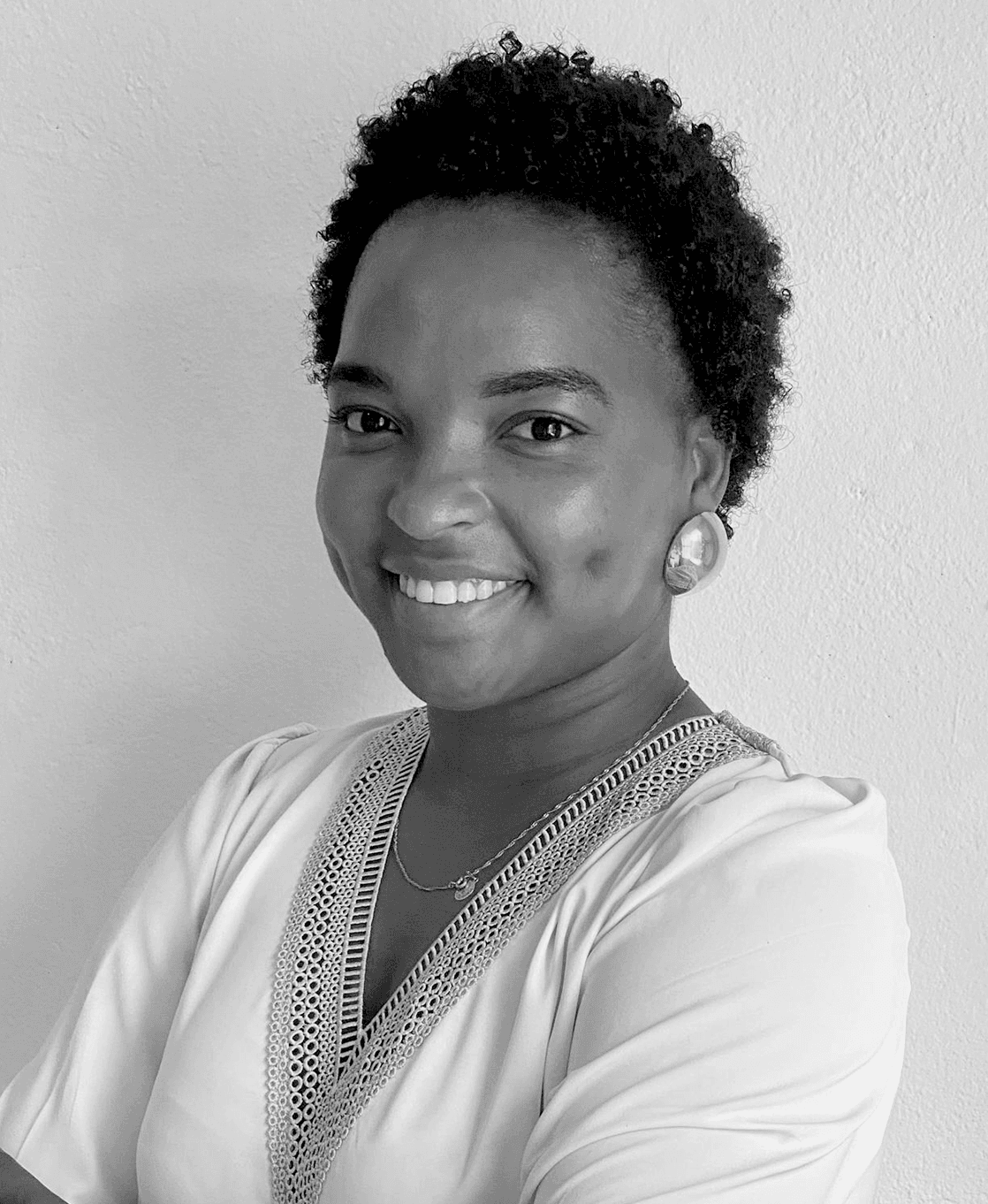 Portrait of Niela Rodrigues in a white blouse with embellishments on the edges, smiling at the camera in front of a white wall