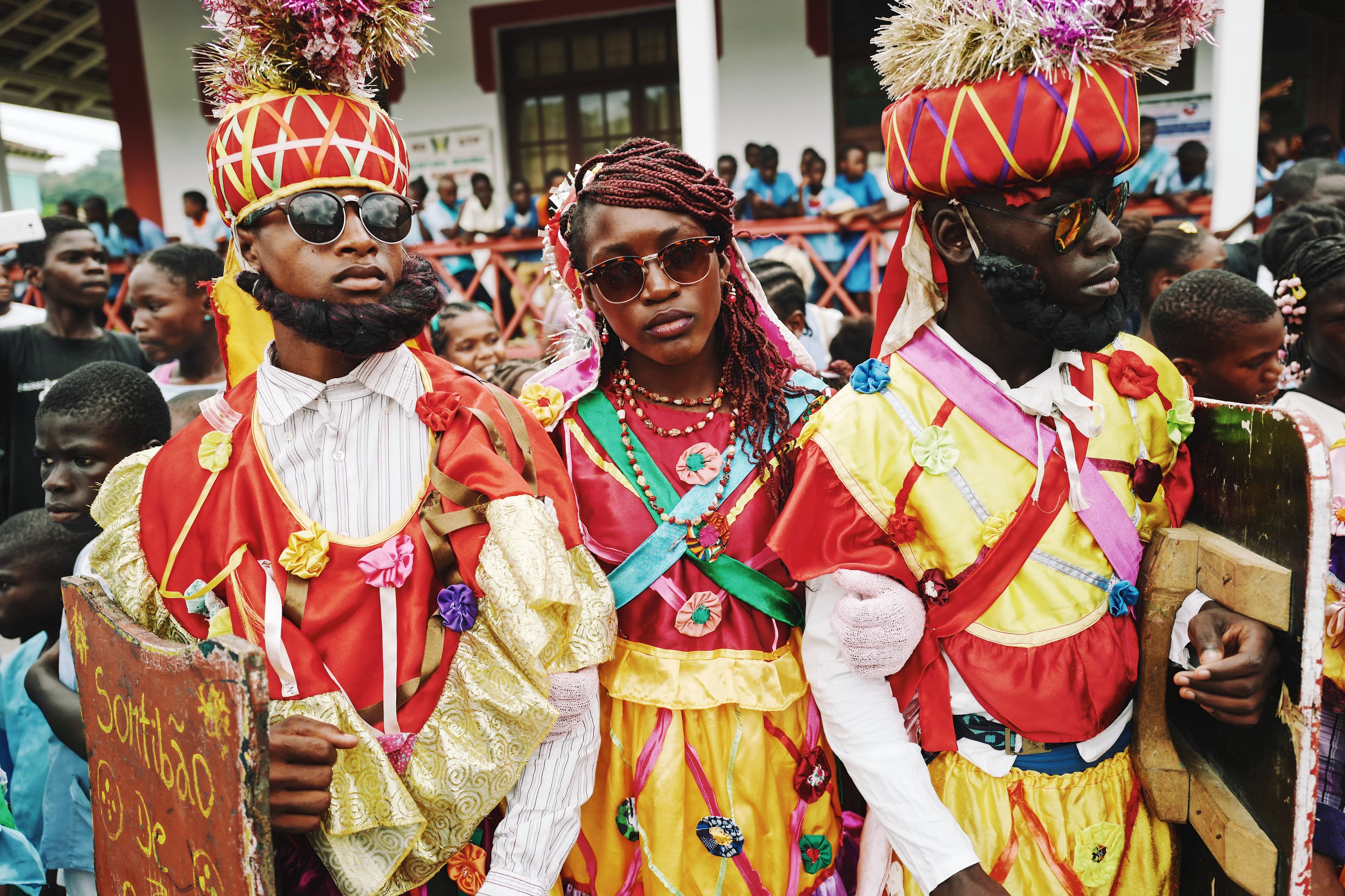 Two men, with a woman in colourful traditional clothes and yellow and red hats, wearing sunglasses in the middle