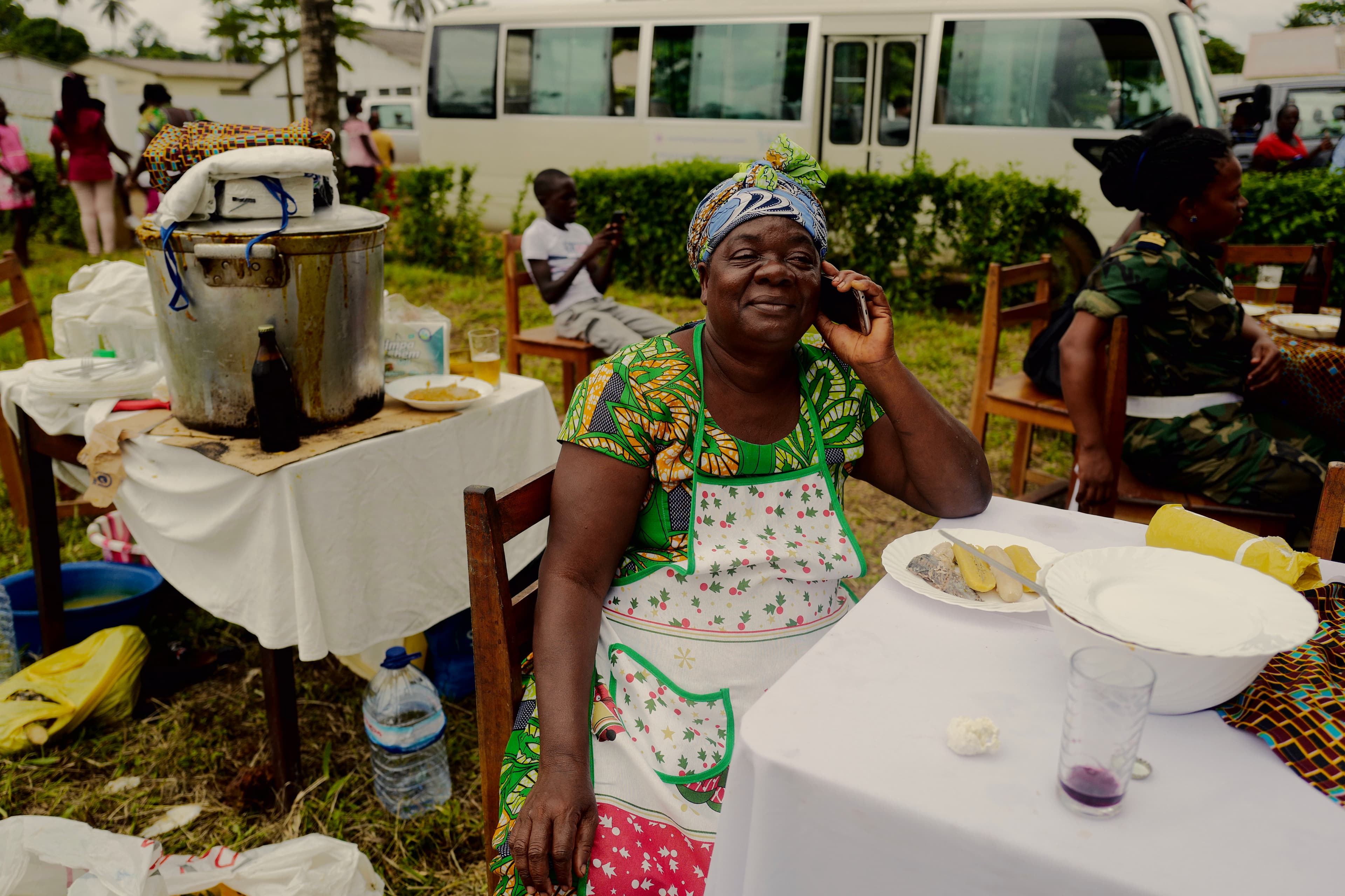 Woman in a colourful dress and apron sits at a set table, talking on the phone